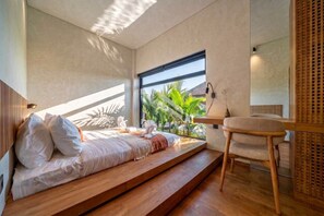 Ein Schlafzimmer mit einem Bett auf einer Holzbodenplattform, einem Nachttisch und Blick auf Grünfläche draußen.