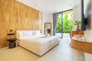 A modern bedroom with a large bed, a wooden headboard, a bedside table, a built-in TV, and a view of lush greenery through a sliding door. - Modern 2BR in Ungasan's Best w Beach Access+Pool (Badung Regency)
