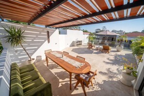 Een overdekt terras buiten, met een eettafel en stoelen, een bank en potplanten.