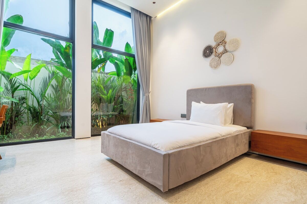 A modern bedroom with a large bed, a view of greenery through floor-to-ceiling windows, and a decorative wall piece.