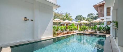 Une piscine entourée de verdure et d’un bâtiment blanc, avec des chaises longues et des parasols.