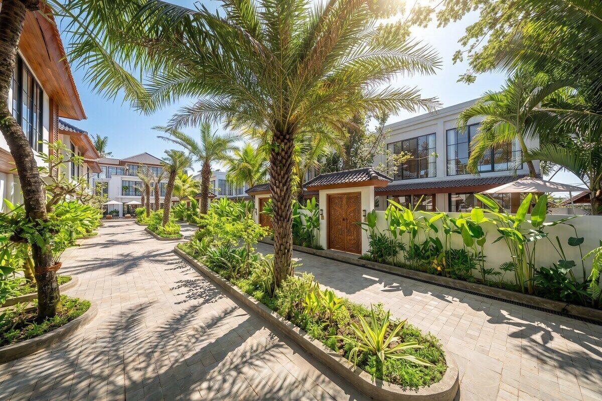 A pathway lined with palm trees and tropical plants leading to a building with a traditional architectural style.