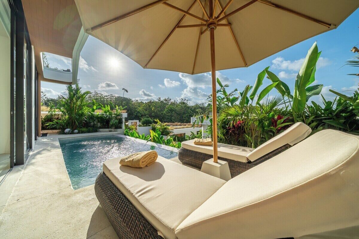 Un área junto a la piscina con sillones de descanso, una sombrilla y abundante vegetación.