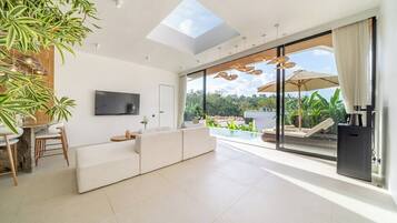 Una sala de estar moderna con televisor de pantalla plana, un sofá, un comedor con sillas y un amplio ventanal que da a una zona exterior con piscina.