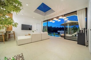 A modern living room with a sofa, a dining area, and a large sliding glass door leading to a pool. - Brand New Private 2BR Villa+Huge Pool+Beach Access (South Kuta)