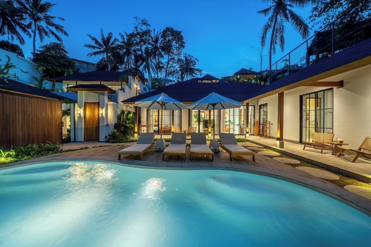 A pool area with lounge chairs and umbrellas, surrounded by modern buildings with large windows and a wooden fence.