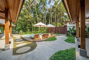Un área de piscina con sillas de descanso y sombrillas, rodeada por una pérgola de madera y palmeras.