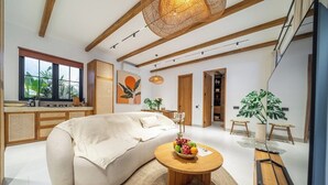 Ett modernt vardagsrum med en soffa, ett runt soffbord i trä med frukt på, en hängande lampa och ett stort fönster med utsikt över grönområden.