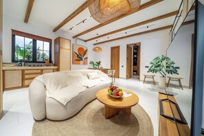 Ein modernes Wohnzimmer mit einer Couch, einem runden Holztisch mit Obst, einer Hängeleuchte und einem großen Fenster mit Blick auf Grünflächen.