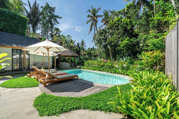 Ein Pool mit Liegestühlen und einer Holzterrasse, umgeben von üppiger Vegetation und Palmen.