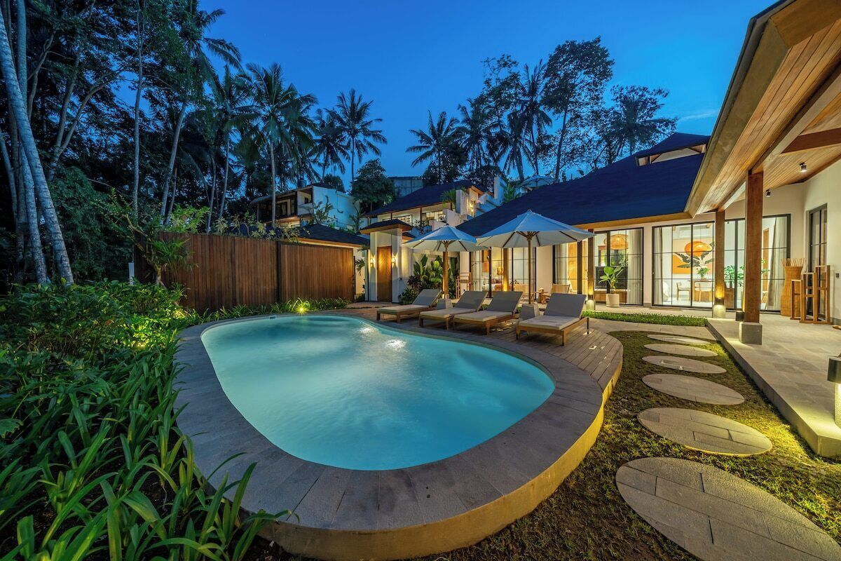 A modern pool area with lounge chairs and umbrellas, surrounded by tropical vegetation and a wooden fence.