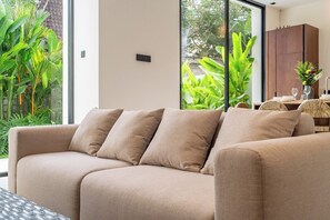 Ein beiges Sofa mit Kissen, ein Esstisch mit Stühlen und Blick auf Grün hinter großen Fenstern.