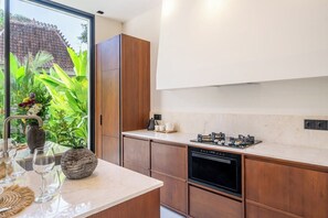 A modern kitchen with wooden cabinets, a built-in oven, and a large window overlooking greenery. - Lily Villa 5 - Cozy 2BR Villa in Serene Canggu w Pool & Garden (Badung Regency)