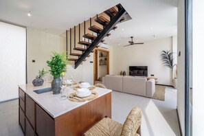 A modern kitchen with a central island, a staircase, and a living area with a sofa and TV. - Lily Villa 5 - Cozy 2BR Villa in Serene Canggu w Pool & Garden (Badung Regency)