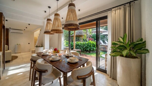 Uma área de refeições com uma mesa de madeira posta para quatro pessoas, cercada por cadeiras, com luminárias de vime pendentes e uma grande planta em vaso.