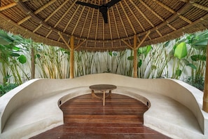 A covered outdoor seating area with a curved bench and a table, surrounded by tall plants and a bamboo ceiling. - Cozy 3BR Villa in Ubud w Garden Bath, Pool & Patio (Gianyar Regency)
