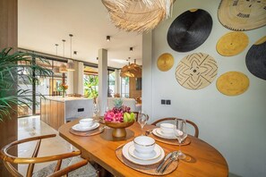 A dining area with a wooden table set for four, a fruit bowl centerpiece, and a wall decorated with circular woven patterns. - Serene 2BR Villa in Ubud w Private Pool & Garden (Tegallalang)