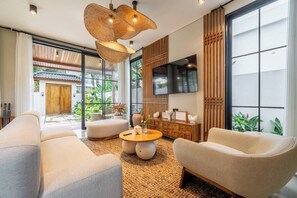 A modern living room with a sofa, armchair, and coffee table, a TV mounted on the wall, and a large window with a view of greenery. - Serene 2BR Villa in Ubud w Private Pool & Garden (Tegallalang)