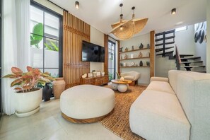 A modern living room with a flat-screen TV, a sofa, a round ottoman, and a staircase with a railing. - Serene 2BR Villa in Ubud w Private Pool & Garden (Tegallalang)