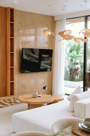 A modern living room with a flat-screen TV mounted on a wooden wall, a round wooden table with a vase, and a sofa with white cushions. - Minimalist 3BR Villa w Pool+Beach Access • Ungasan (Badung Regency)