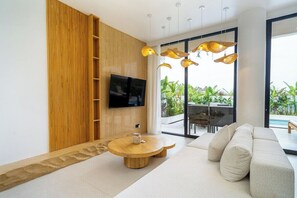 A modern living room with a flat-screen TV, a round wooden coffee table, a beige sofa, and a large window with a view of greenery. - Minimalist 3BR Villa w Pool+Beach Access • Ungasan (Badung Regency)