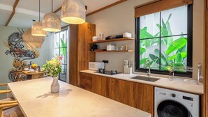 A modern kitchen with wooden cabinets, a built-in washing machine, and a large window with a view of greenery. - Tropical 2BR Villa w Private Pool & Garden • Ubud (Gianyar Regency)