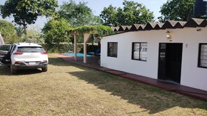 A white building with a covered outdoor area, a car parked in front, and a pool in the background.
