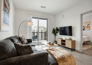 Inviting living area with a plush sectional, stylish décor, and a large wall-mounted TV. Sliding doors open to a private balcony, while the adjacent bedroom offers added convenience and flow. - Modern Apt with Pool, Garage & Laundry (Madison)