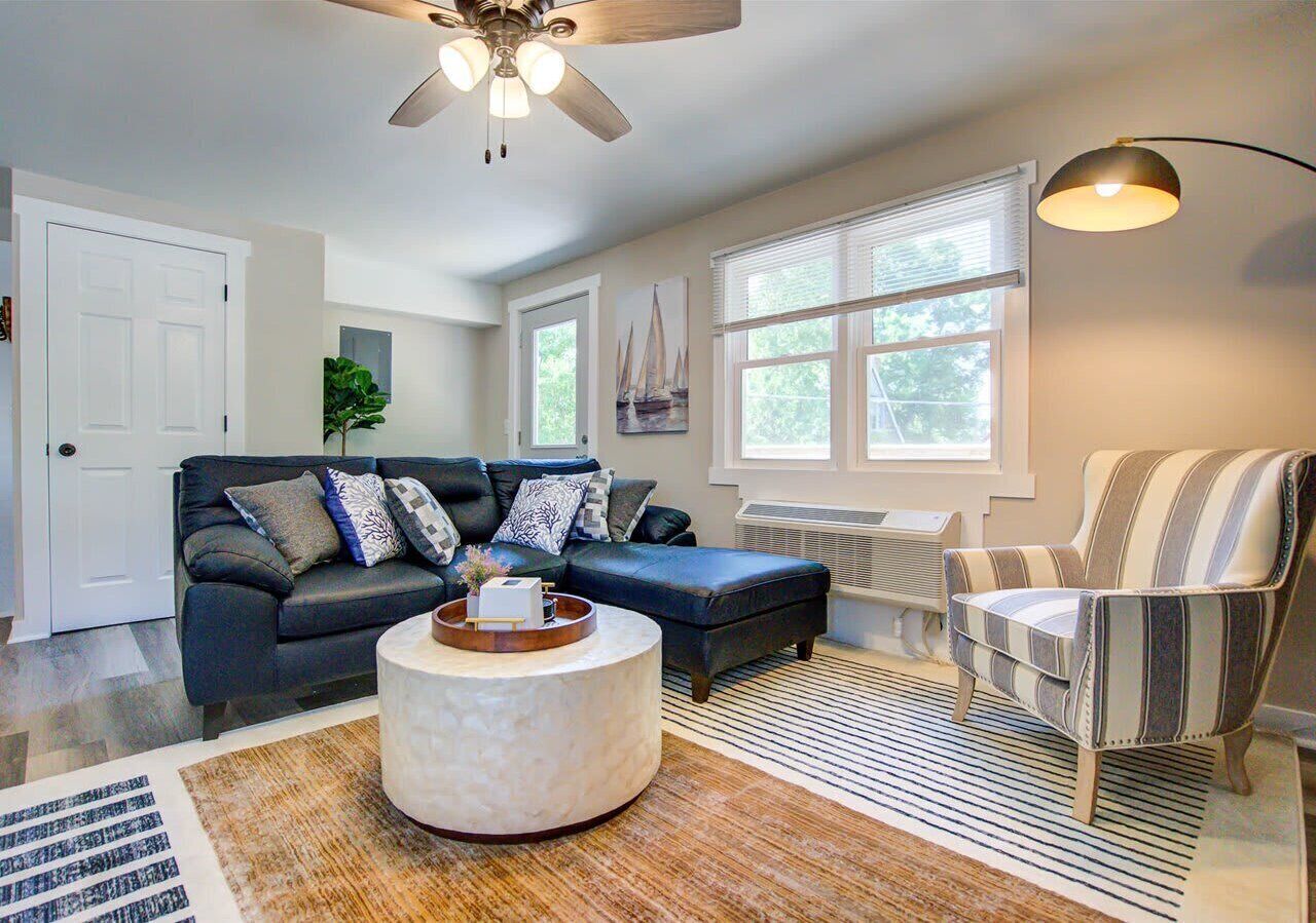 Bright living area with a stylish sectional, accent chair, and round coffee table. Large windows bring in natural light, creating a welcoming space to relax or entertain.