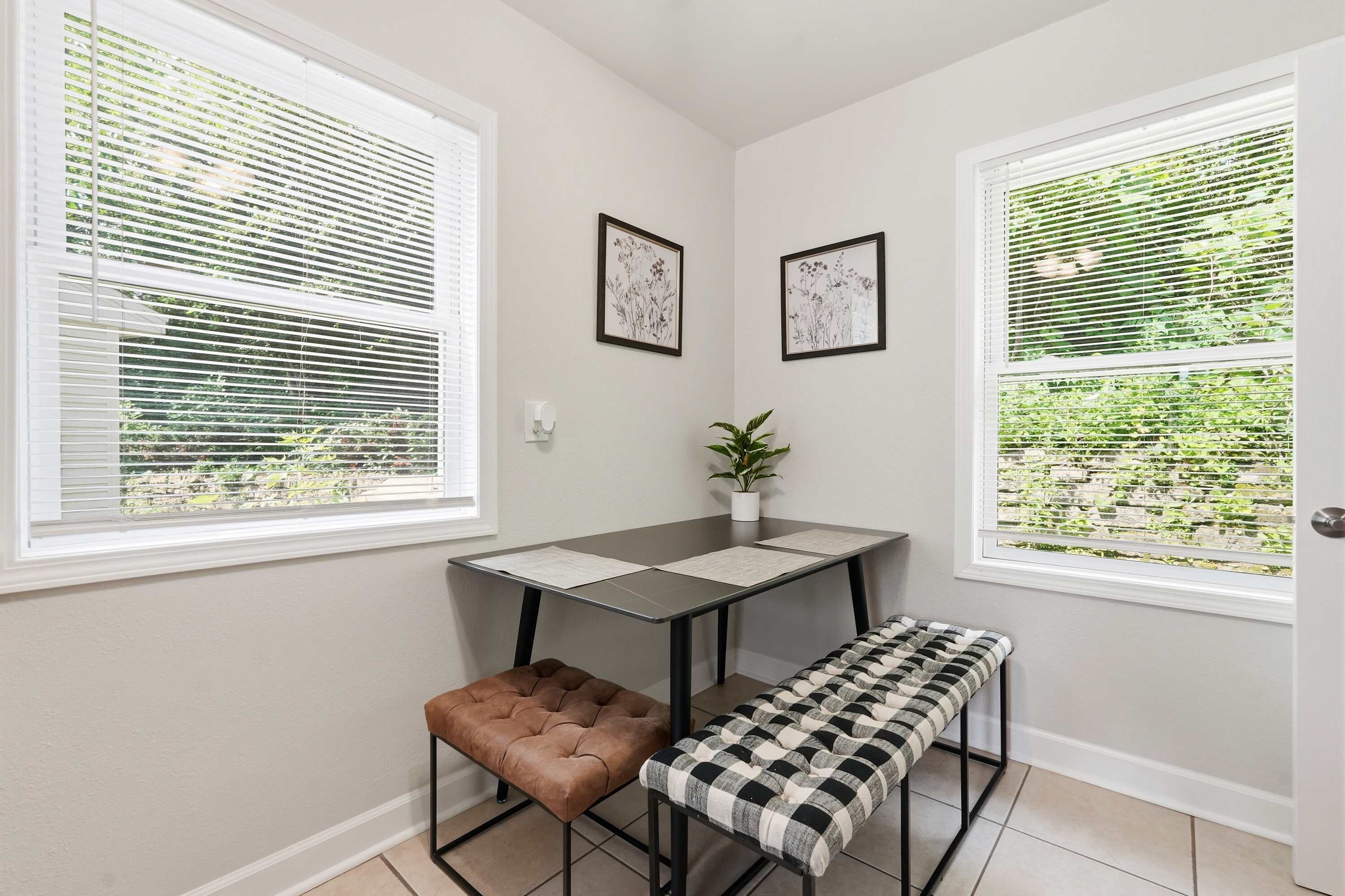 Une pièce d’angle avec deux fenêtres, une table, un banc et deux tableaux encadrés au mur.