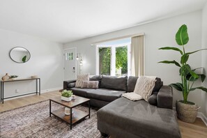Ein modernes Wohnzimmer mit einem grauen Sofa, einem Couchtisch, einer großen Kübelpflanze und Blick auf Grünflächen.