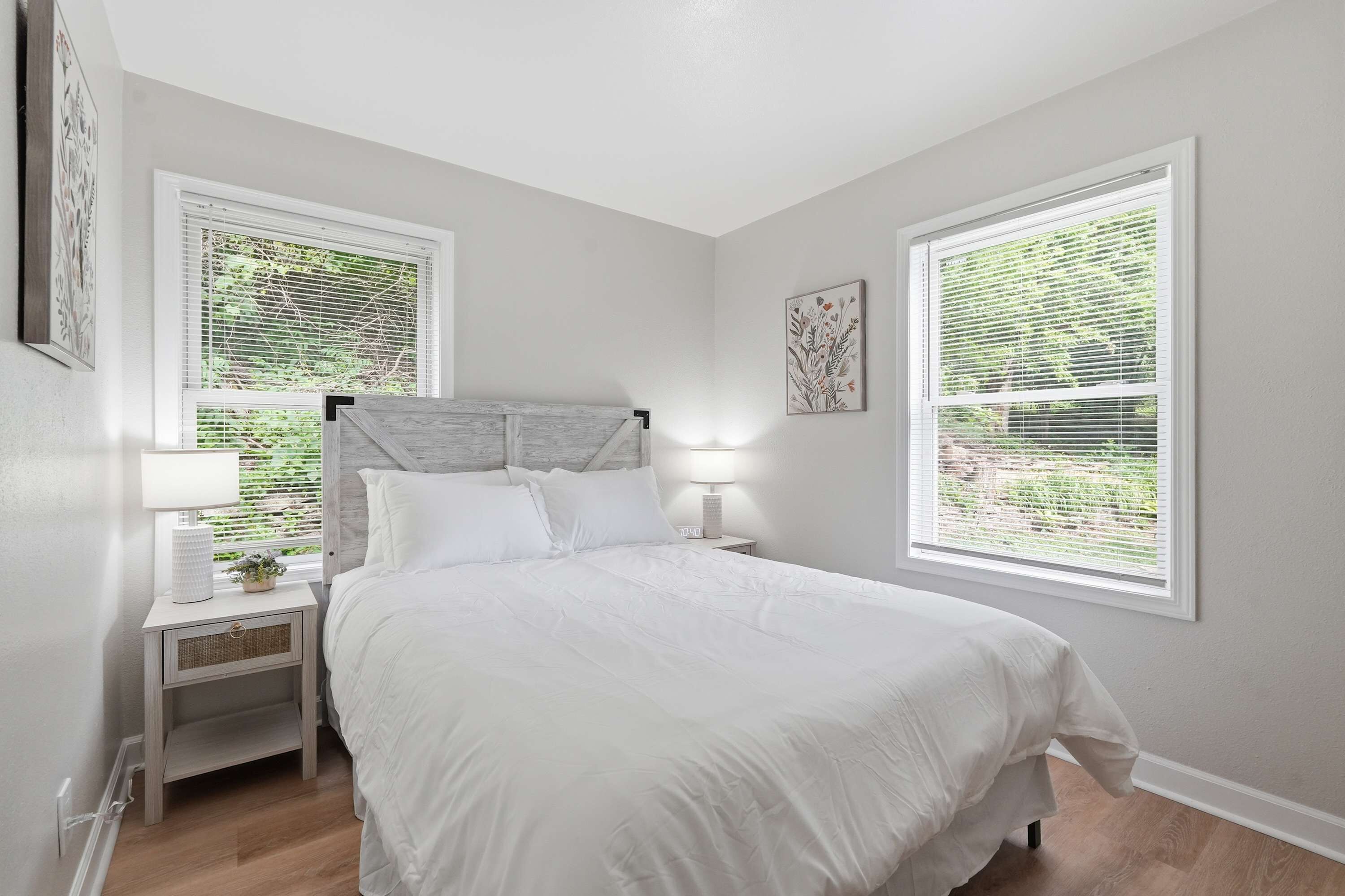 A bedroom with a bed, two bedside tables, a nightstand, a wall-mounted lamp, and two windows with blinds.