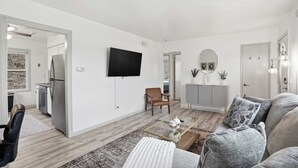 Ett modernt vardagsrum med en platt-TV, en soffa, ett soffbord och ett köksområde som syns genom en öppen dörr.
