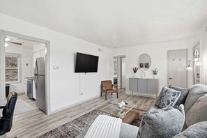 Ein modernes Wohnzimmer mit Flachbildfernseher, einer Couch, einem Couchtisch und einem durch eine offene Tür einsehbarer Küchenbereich.