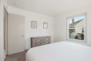 Ein Schlafzimmer mit einem Bett, einem Schminktisch, zwei gerahmten Bildern und einem Fenster mit Blick auf ein Haus.