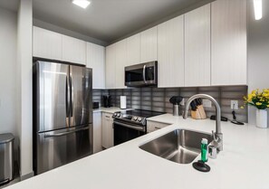 Contemporary kitchen with stainless steel appliances, a sleek tile backsplash, and crisp white cabinetry. The spacious island and modern fixtures create a stylish and functional space for cooking or entertaining. - Poolside Apt with Garage & In-Unit Laundry (Madison)