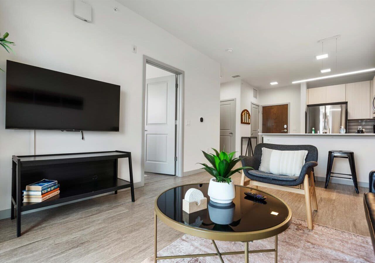 Bright living room with wall-mounted flat-screen TV and modern black console. A round coffee table with a potted plant centers the space, while neutral floors and open sightlines to the sleek kitchen create an airy, contemporary feel.