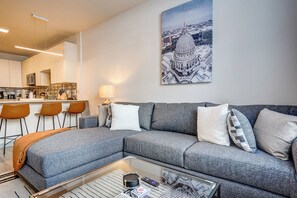 Stylish open-concept living room with a plush gray sectional, accent pillows, and a cozy throw. A sleek glass coffee table faces the kitchen with bar seating, creating a welcoming space to relax or entertain. - Modern Apt with Pool, Parking & Pet Wash (Madison)