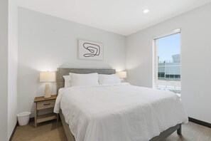 Ein modernes Schlafzimmer mit einem großen Bett, zwei Nachttischlampen, einem Nachttisch, einer kleinen Mülltonne und einem an der Wand hängenden, abstrakten Kunstwerk.