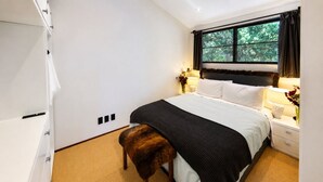 Ein Schlafzimmer mit einem Bett, Nachttischen, einem Fenster mit Vorhängen und einem kleinen Hocker.