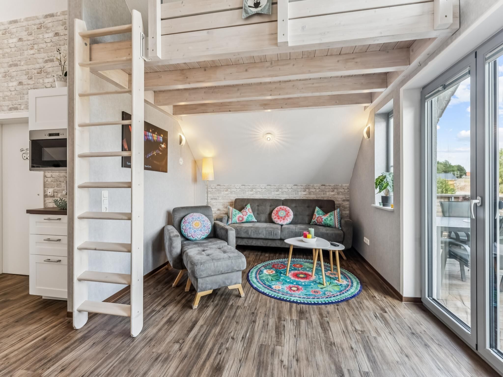 Un espace de vie de style loft avec un lit en mezzanine, un canapé gris agrémenté de coussins colorés, une petite table ronde et une grande fenêtre donnant sur l’extérieur.