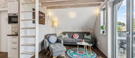 Un espace de vie de style loft avec un lit en mezzanine, un canapé gris agrémenté de coussins colorés, une petite table ronde et une grande fenêtre donnant sur l’extérieur.
