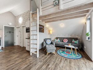 Ein Loft-Wohnraum mit Schlafgalerie, einem grauen Sofa mit gemusterten Kissen, einem runden Couchtisch und Holzboden.