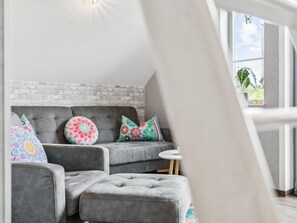 Uma sala de estar moderna com um sofá cinza, almofadas coloridas, uma mesa redonda pequena e uma grande janela com vista para o verde.