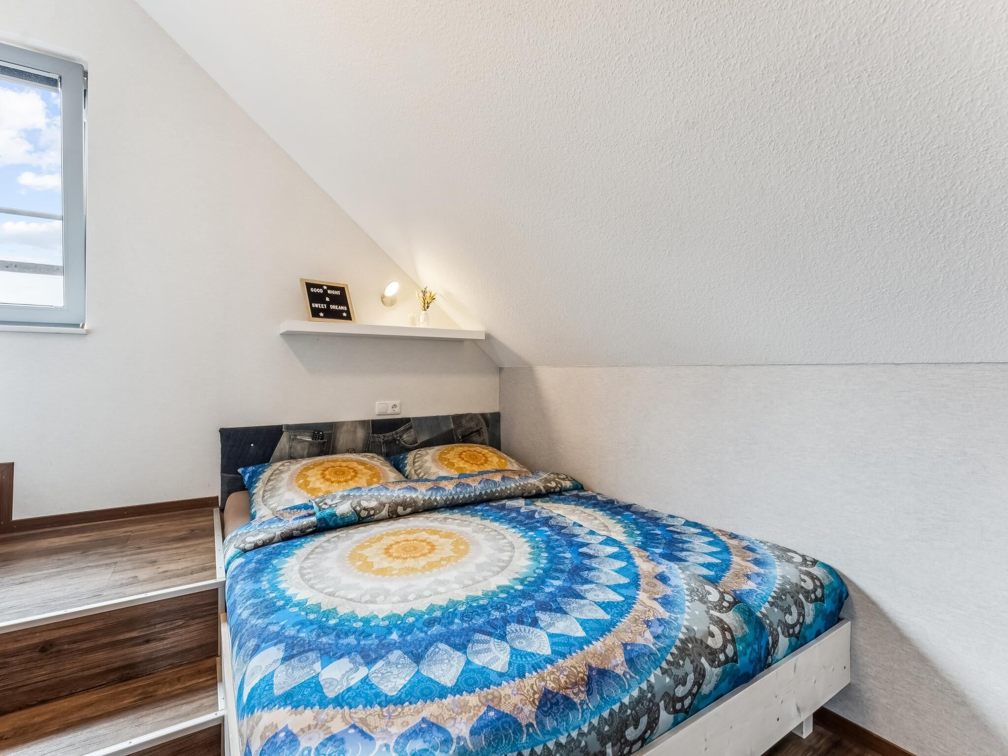 A bedroom with a bed, a nightstand, a shelf with a sign, and a window.