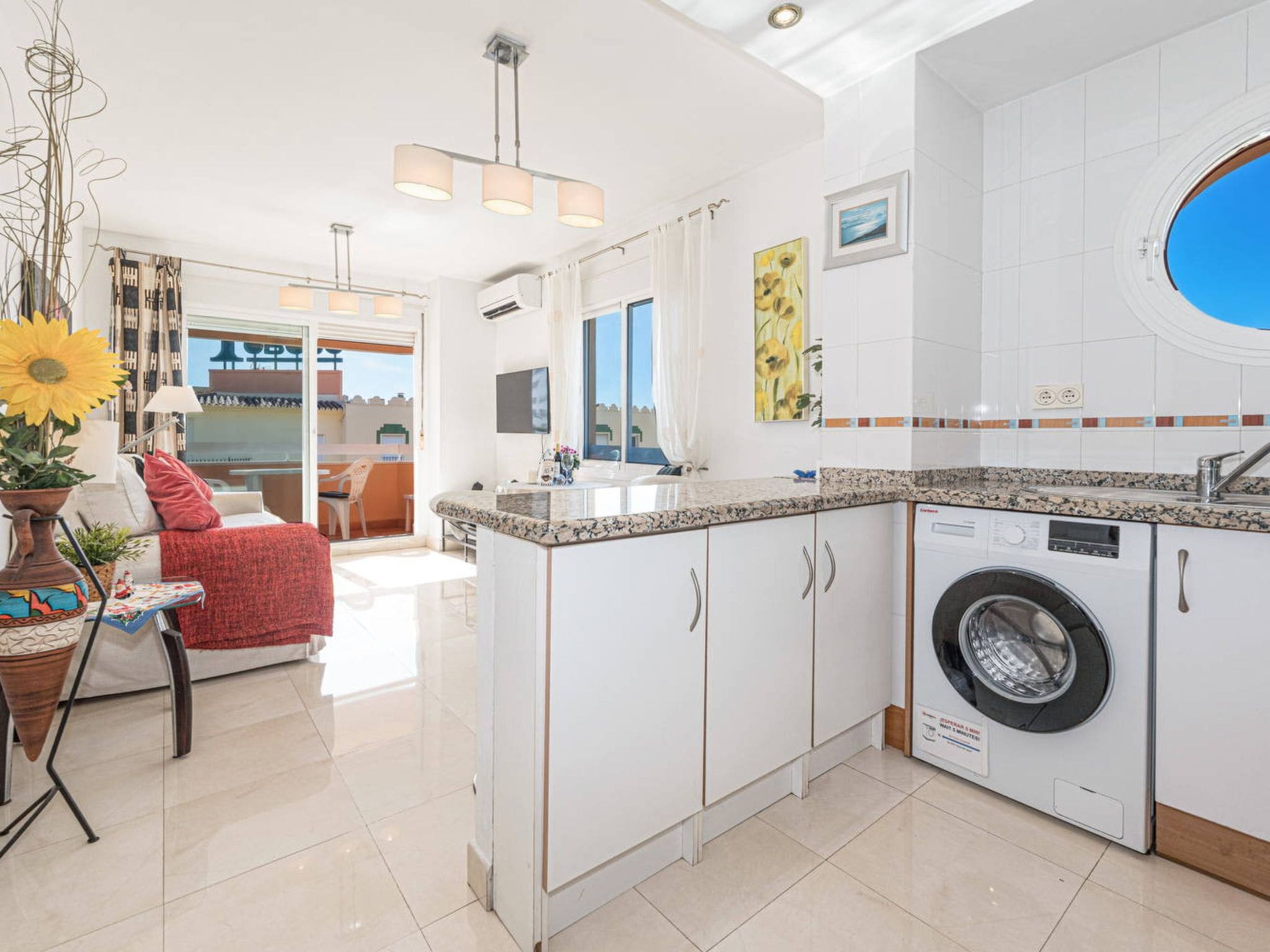 A compact kitchen with a built-in washing machine, a dining area with a red sofa, and a view of the outside.