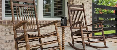 Two wooden rocking chairs with a small table between them, positioned in front of a brick house with large windows.