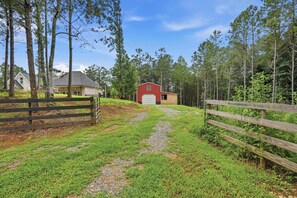 A rural property with a gravel path leading to a red barn, surrounded by tall trees and a wooden fence.