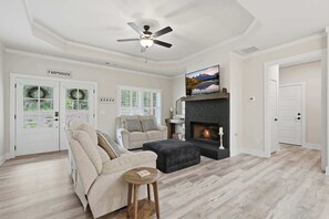 A spacious living room with a fireplace, a ceiling fan, and a large window with a view of the outdoors.