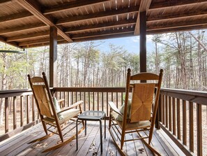 Eine Holzterrasse mit zwei Schaukelstühlen und einem kleinen Tisch, mit Blick auf ein bewaldetes Gebiet.